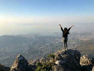 Une jeune fille se tient au sommet d’une montagne en Afrique du Sud, les bras levés en signe de victoire, admirant une vue spectaculaire sur la ville du Cap pendant l’année scolaire en Afrique du Sud.